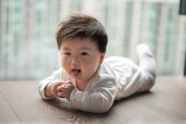 Baby with thick, dark hair lying on tummy looking at camera with hands touching, head raised, and tongue out slightly